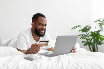 Smiling millennial african american bearded man lies on white bed in bedroom interior shopping online