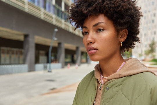 Outdoor shot of lovely thoughtful young woman focused into distance dressed into distane strolls in city thinks about something has walk during spare time enjoys good sunny day during weekend.