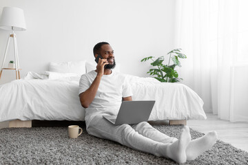 Glad young african american bearded guy sitting by bed working on laptop talking by phone with client