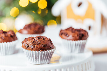 Chocolate cupcakes or muffins with Ginger house and Christmas lights from Christmas tree in blurry background. Festive and holiday decoration