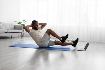 Fototapeta premium Happy young african american guy doing exercises for legs and abs on mat on floor in room interior