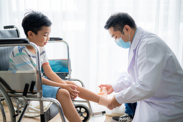 Obraz premium Asian boy sitting on a wheelchair is having the doctor wrap a cloth around his leg because he broke his leg at the hospital.