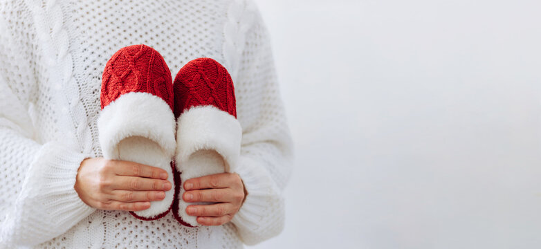 Woman in white sweater holds warm knitted red slippers. Concept of home family cozy holiday Christmas New Year.