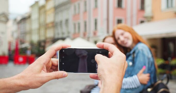 Beautiful redheaded girl and her female friend posing for camera in city. Guy holding smartphone and taking pictures of women hugging and smiling. Technology, travel, social media concept.