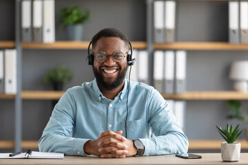 Smiling millennial african american bearded guy in glasses and headset working on laptop and looking at camera