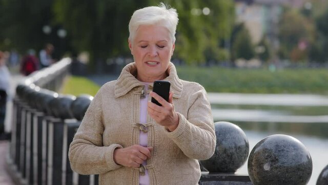 Happy senior woman of retirement age making video call, looking at smartphone camera. Attractive old mature female uses modern gadget wireless internet tell good news talking on mobile phone outdoor