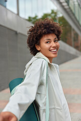 Vertical shot of happy carefree sporty woman smiles gladfully carries fitness mat dressed in anorak...