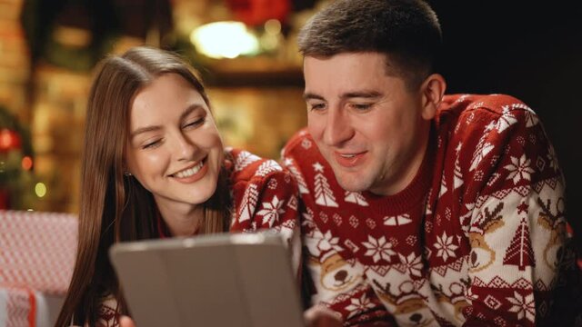 Happy caucasian couple lying home near Christmas tree background. Beautiful woman with long hair holds digital tablet and emotionally tells funny stories