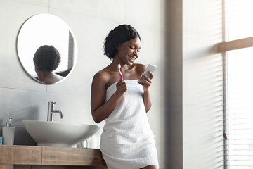 Black woman using smartphone while brushing teeth in bathroom