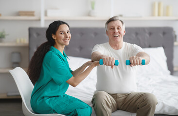 Female physiotherapist helping positive mature man to do exercises with dumbbells on bed at retirement home