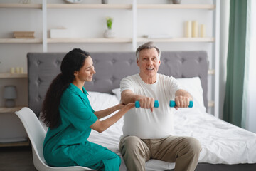 Young physiotherapist helping senior man to do physical exercises with dumbbells on bed at home - obrazy, fototapety, plakaty
