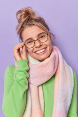 Indoor shot of lovely young fair haired woman tilts head keeps hand on rim of spectacles smiles broadly looks joyfully at camera hears good news wears long sleeved jumper and warm scarf around neck