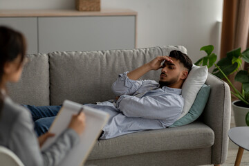 Fototapeta premium Young arab male patient lying on couch at psychologist's office, having session with counselor, guy feeling depressed