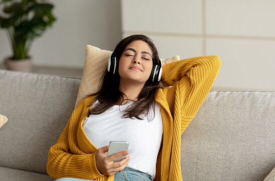 Young relaxed arab woman in wireless headphones relaxing on couch, listening to music at home, free space
