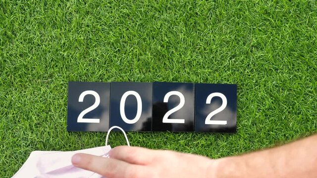 Man hand changes plastic plates with numbers from 2021 to 2022 on green grass with fabric face masks, takes masks and throws them away. Concept of the end of global pandemic situation, free of covid