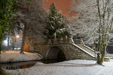 Kamienny mostek nad rzeką w parku dworskim w mieście Iłowa, w Polsce. Na ziemi i gałęziach drzew leży warstwa śniegu. Jest noc.
