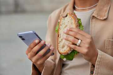 Cropped shot of unrecognizable woman eats delicious sandwich and uses mobile phone for texting...
