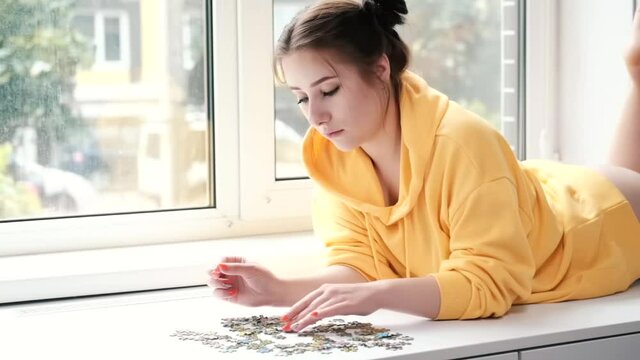 pretty brunette girl in yellow hoodie arranging pieces of a Jigsaw Puzzle Game in bright white room. playing board game home