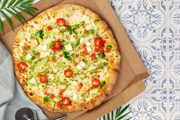 Natural italian big pesto pizza with thin dough, chicken, mozzarella, cherry tomatoes and bazil in a craft box on a tile table with palm leaves. Mediterranean cuisine. Healthy and testy fast food