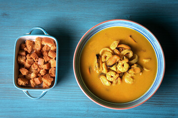 Seafood soup, based on prawns, mussels, clams, hake and squid, accompanied with pieces of fried bread, on a light blue wooden table.