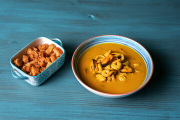 Seafood soup, based on prawns, mussels, clams, hake and squid, accompanied with pieces of fried bread, on a light blue wooden table.