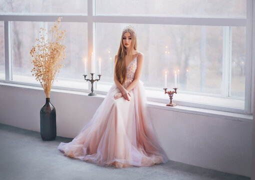 fantasy medieval girl princess in sitting on windowsill of large white window classic room. Woman queen beauty face. Vintage glamour dress golden luxury crown, long loose blonde hair. Fashion model
