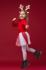 happy funny little girl in Golden reindeer antlers running and jumping on red background in Studio on Christmas day. The child runs and jumps in full growth. Dynamic image. Copy space.