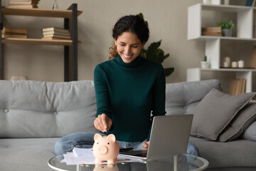 Smiling young Hispanic woman put coin into piggybank save money for future, pay bills taxes online...