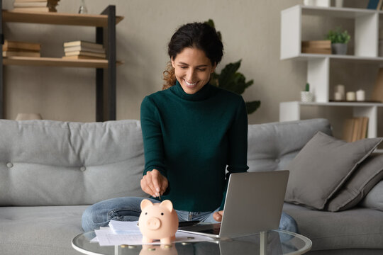 Smiling young Hispanic woman put coin into piggybank save money for future, pay bills taxes online on computer. Happy Latin female manage household budget finances, make investment in piggy bank.