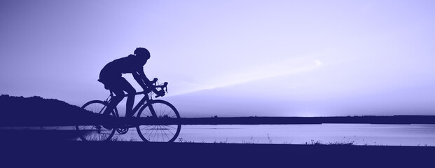 Unrecognizable silhouette man riding bicycle against sunset sky. Road biking cyclist workout, riding racing bicycle on open road. Toned in trendy Color Of The Year 2022 Very Peri.. Copy space, banner