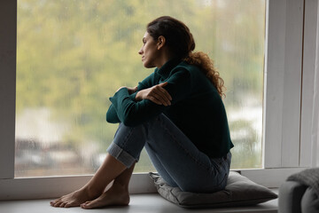 Pensive sad millennial Hispanic woman sit on windowsill at home look in window distance thinking...