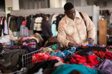 Fotobehang african american guy chooses sacond hands clothes at flea market © caftor