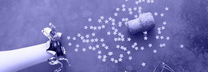 Champagne bottle with confetti stars on dark grey background. New year, birthday, anniversary, or wedding concept. Flat lay, banner. Toned in trendy Color Of The Year 2022 - Very Peri.