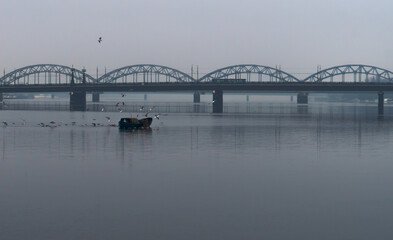 Naklejka premium A little wooden fishing boat in the calm river who flow through big city. In the background stone bridge with traffic on it. Winter time.