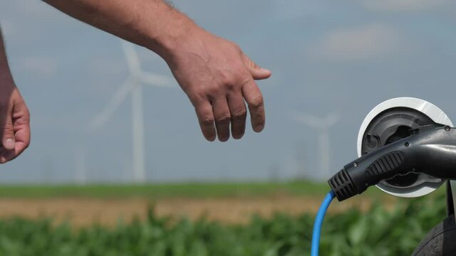 Wind turbines rotate in corn field. Focus changes from powerful operating machines to man connecting plug to e-car for charging at station closeup