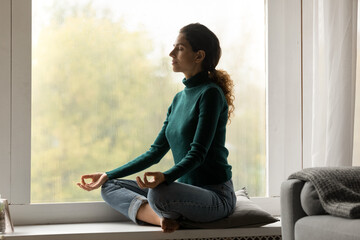 Peaceful Hispanic woman sit on windowsill at home practice yoga with mudra hands breathe fresh air....