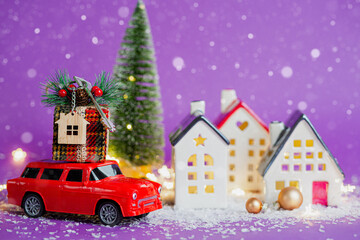 Red retro car on snow carries gift box with home key with keychain cottage on roof past houses with fairy lights, Christmas tree. Violet background. Cozy New Year. Real estate, relocation, mortgage.