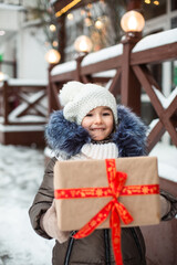 Portrait of joyful girl with a gift box for Christmas on a city street in winter with snow on a festive market with decorations and  fairy lights. Warm clothes, knitted hat, scarf and fur. New year