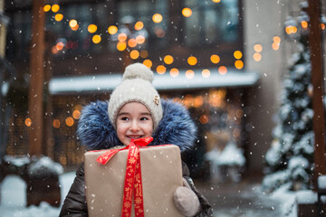 Portrait of joyful girl with a gift box for Christmas on a city street in winter with snow on a festive market with decorations and  fairy lights. Warm clothes, knitted hat, scarf and fur. New year