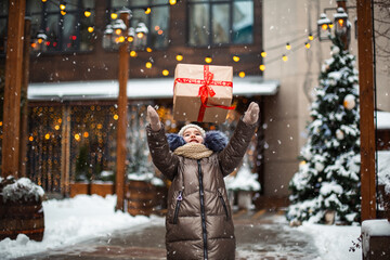 Obraz premium Portrait of joyful girl with a gift box for Christmas on a city street in winter with snow on a festive market with decorations and fairy lights. Warm clothes, knitted hat, scarf and fur. New year