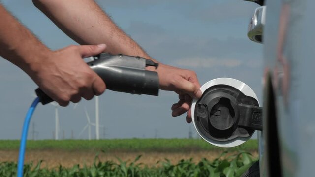 Wind turbines rotate at rural station. Man opens cap and puts plug in white e-car against modern windmills operating in corn field closeup