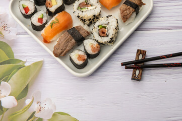 Diverse selbstkreierte Sushivarianten ordentlich Angerichtet auf einen Teller mit weißem Holzuntergrund, Essstäbchen und Blumendekor
