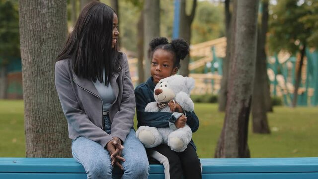 Mom and daughter sitting on bench child holding teddy bear little girl showing five fingers african american family communicates outdoors in park parent smiling listens kid mother speaking with pupil