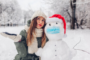 Sad woman sympathizes sick snowman wearing medical mask and Santa hat in snowy winter park. Safety...