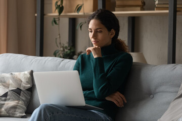 Pensive millennial hispanic woman sit on sofa at home work online on laptop think or make decision. Thoughtful young Latino female look at computer screen plan or solve problem. Technology concept.