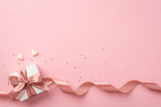 Top view photo of st valentine's day decorations white giftbox with bow pink curly ribbon small hearts and sequins on isolated pastel pink background with empty space