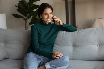 Smiling young Hispanic woman sit on sofa in living room speak talk on cellphone with good mobile provider connection. Happy Latin female have pleasant smartphone call. Communication concept.