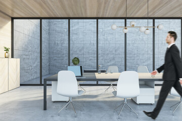 Businessman in suit walking in modern wooden and concrete meeting room interior with panoramic glass windows, table with devices.