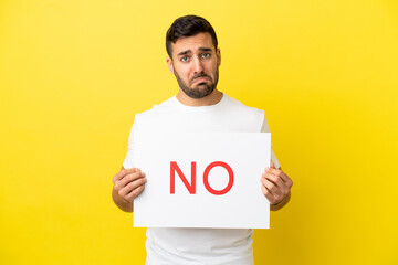Young handsome caucasian man isolated on yellow background holding a placard with text NO with sad expression
