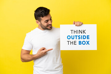Young handsome caucasian man isolated on yellow background holding a placard with text Think Outside The Box and  pointing it